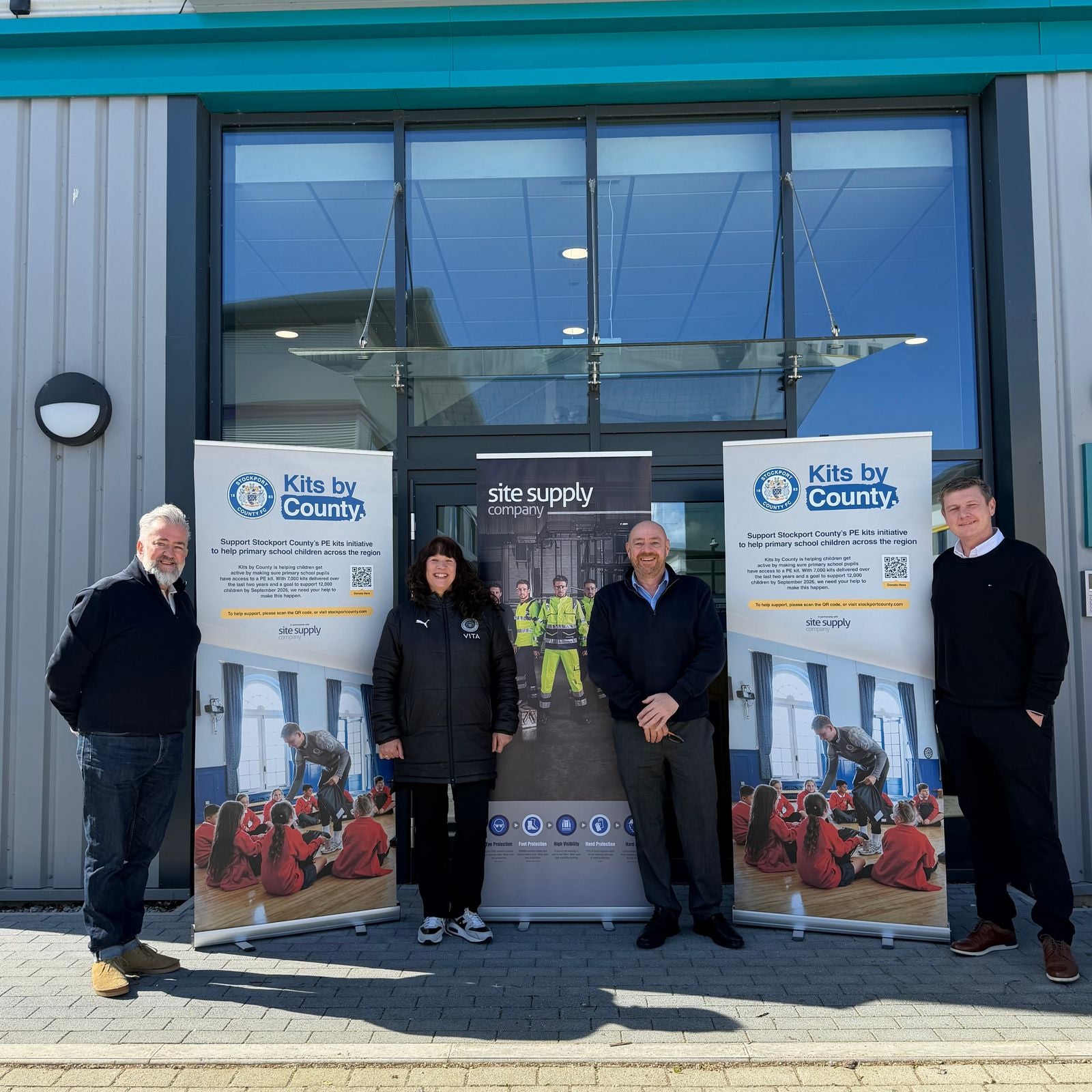Four people with Kits by County and Site Supply Company banners outside the premises